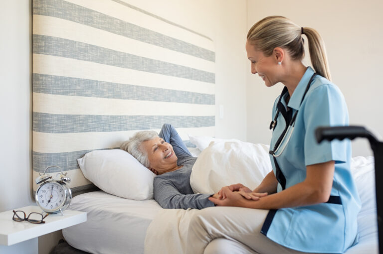 Friendly nurse with senior woman at home. Smiling nurse talking with her patient while getting her out of bed. Disabled woman getting regular health checkup done at home with doctor.